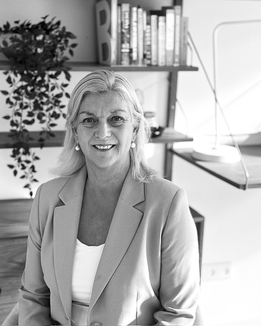 Zwart-wit portret van Hetty Kerkhof, eigenaresse van Zee van Talent en Gallup Certified Strengths Coach, lachend in haar werkruimte.